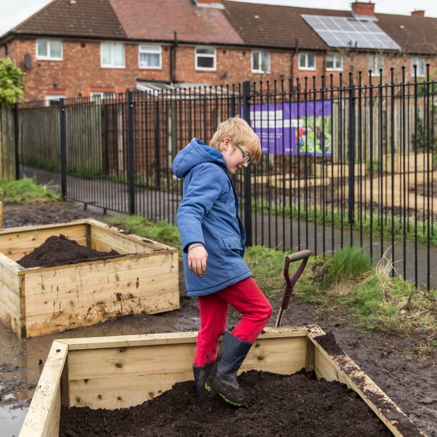 A child in a raincoat and wellies stomps on the soil in a raised planting bed. There is a lane and some houses in the background.