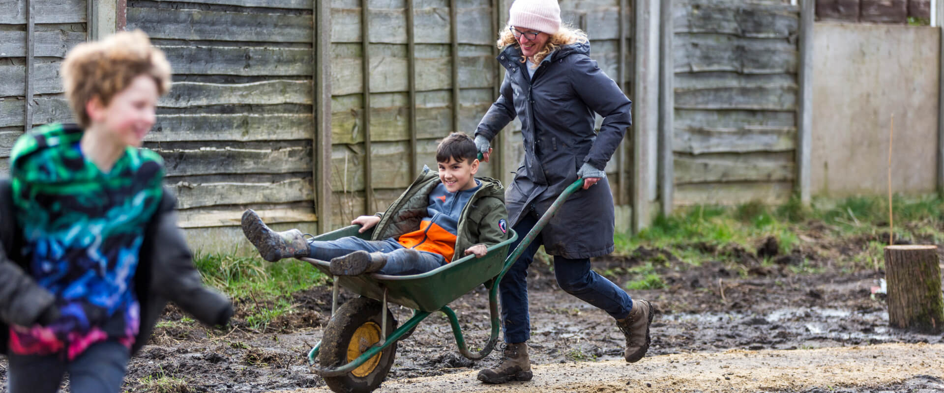 An adult pushing a young person along a path in a wheelbarrow following another young person running. Everyone is smiling.