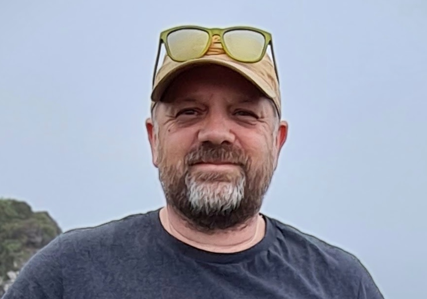 An image of Simon Needle, smiling at the camera. He is wearing a dark blue t-shirt and has sunglasses perched on his head.
