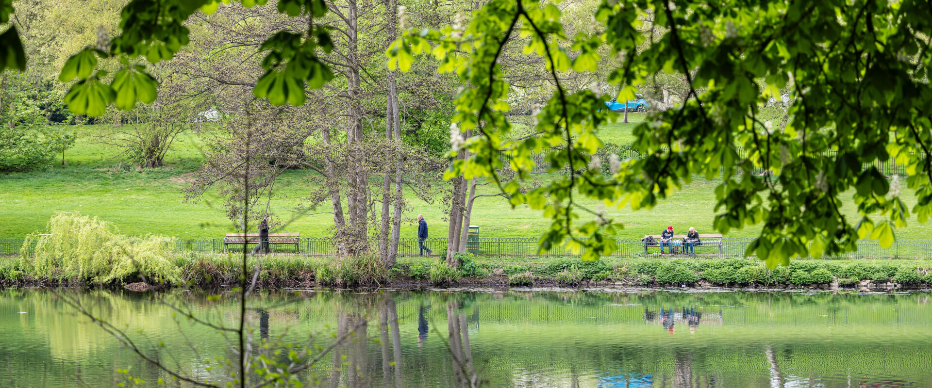 A pond in a park. At the far side people walk along the path and sit on a bench.