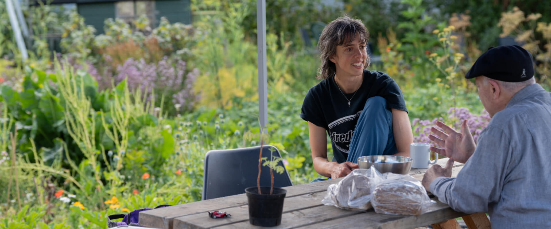 A younger and older person facing each other, sat at a wooden table, smiling while talking within a community garden.