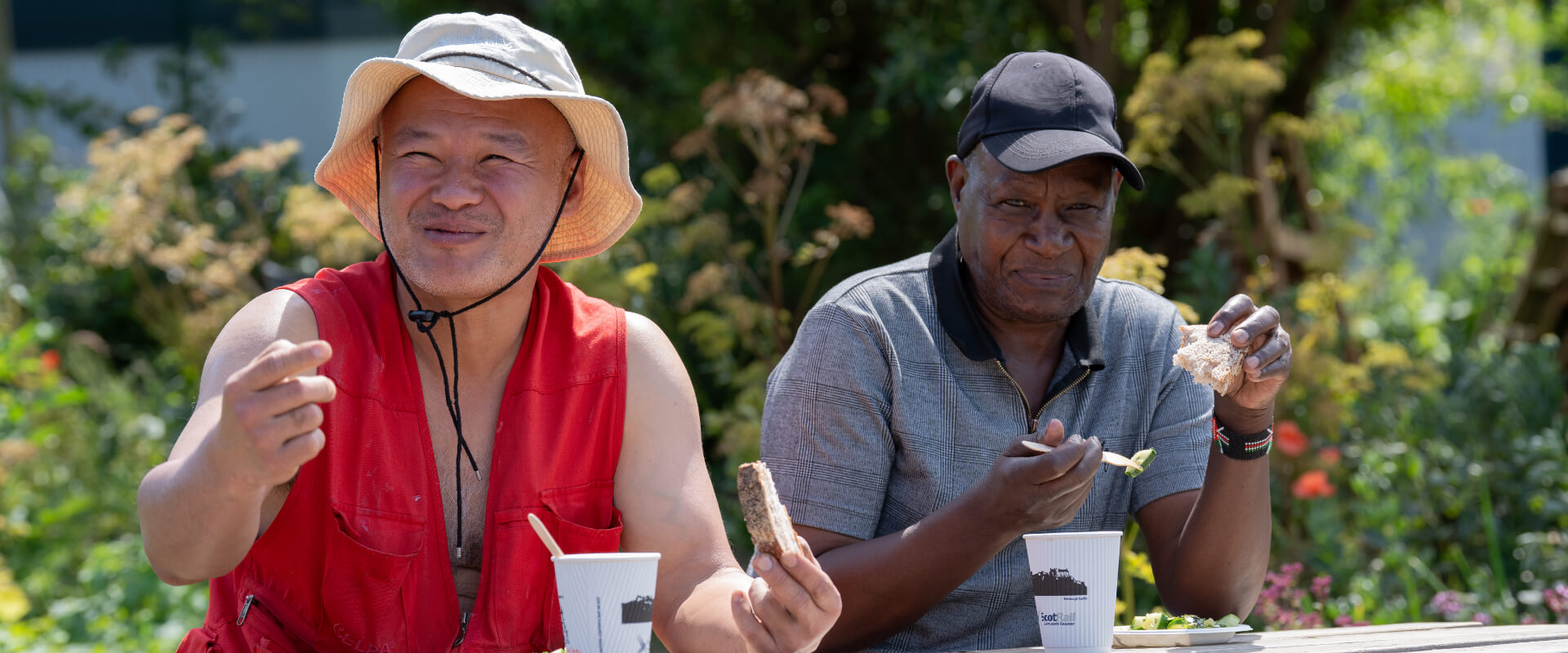 Two men smiling while eating and drinking refreshments in a community garden.
