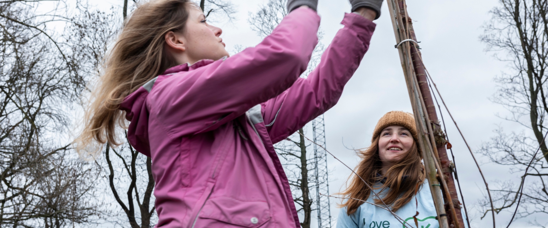 Two girls, one in a pink waterproof jacket and one in light-blue top and yellow hat, smile as they get stuck in with tree planting on a winters' day