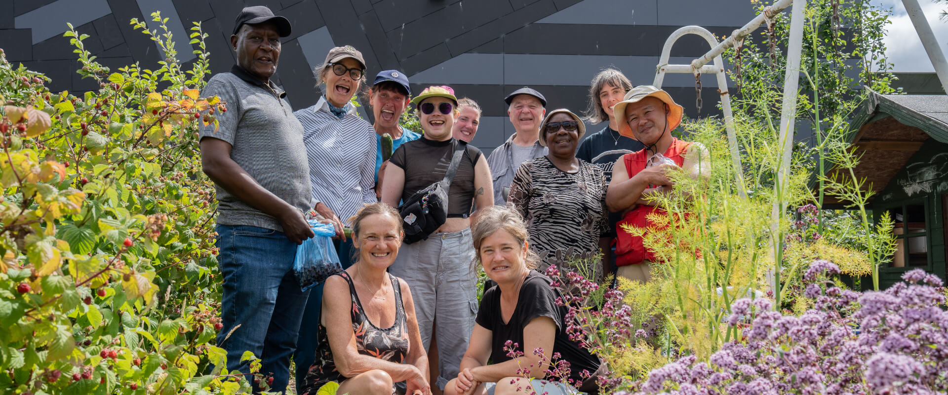 A group of eleven people smiling within a community garden surrounded by flowering plants.
