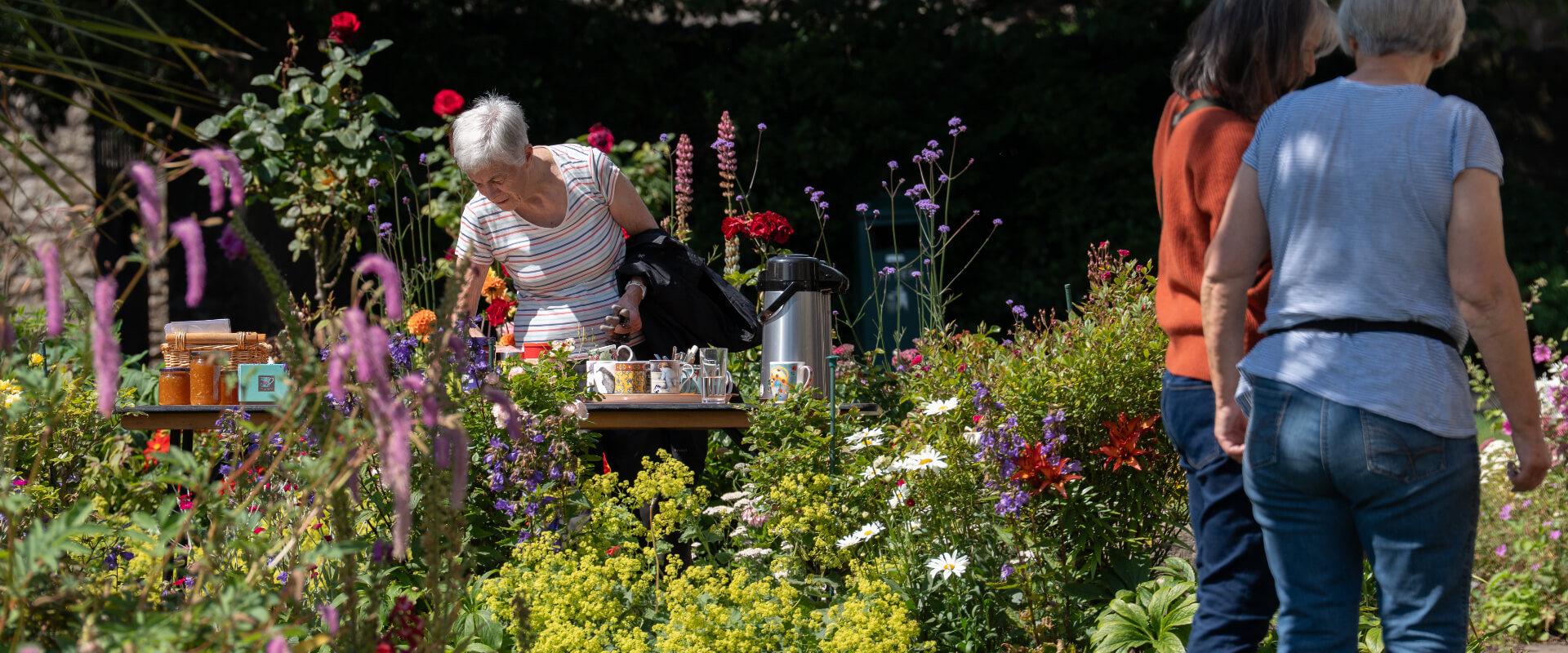 A woman leaning over a refreshments table in a flower garden. Two other people are facing away from the camera in conversation.