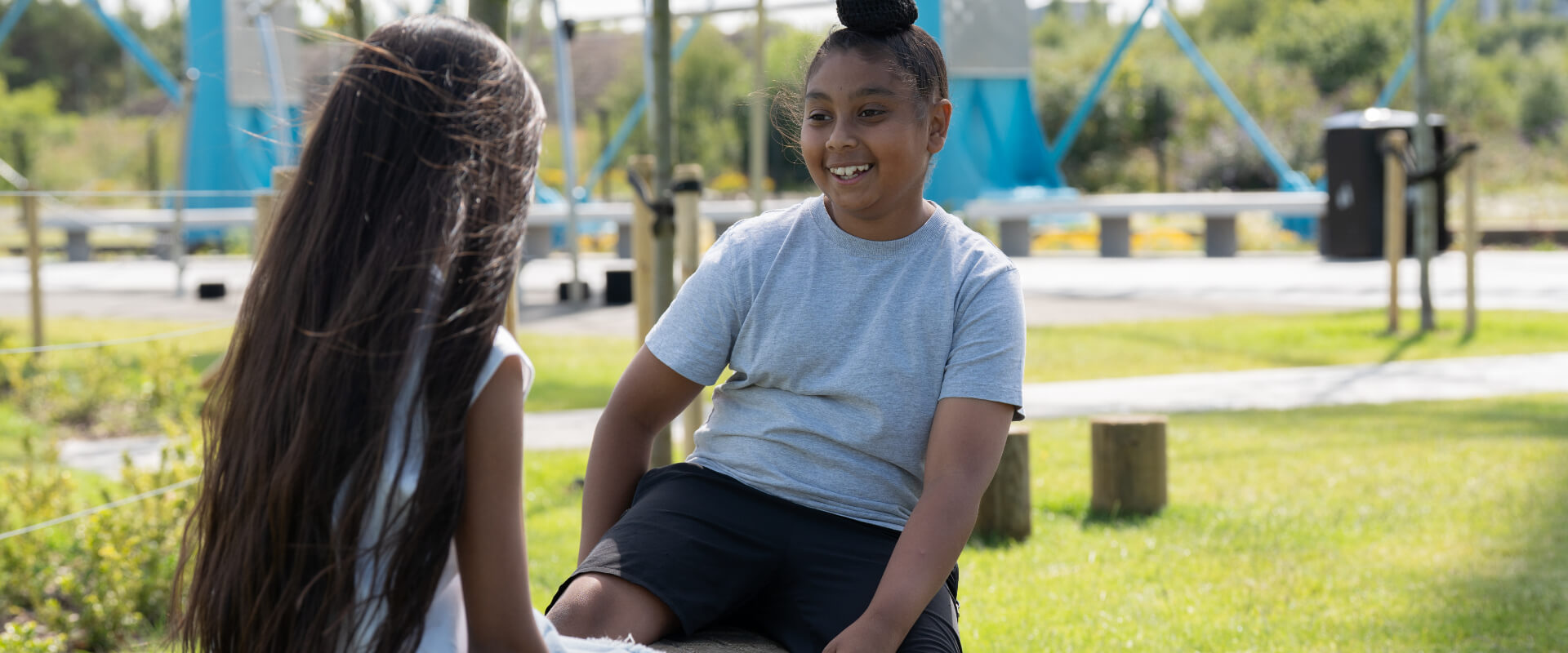 A young person smiling towards another in a park play area.