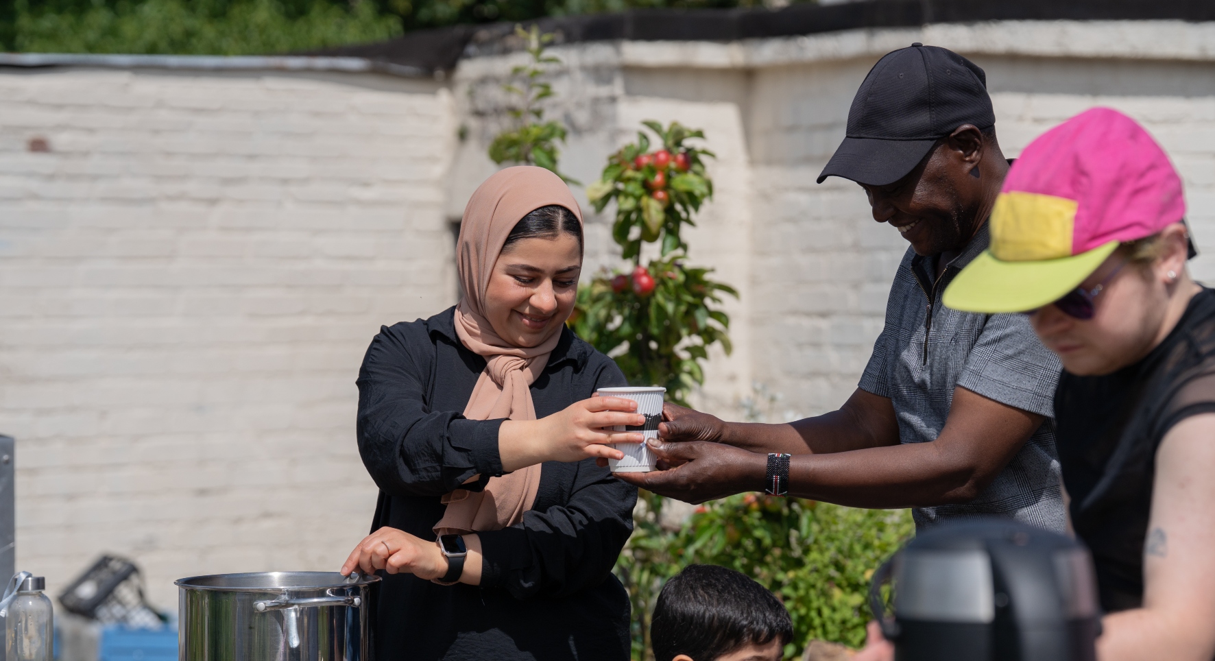 A young lady standing in front of a white garden wall smiles as she passes a cup to a man with outstretched arms. She looks to have ladled the cup contents from a large cooking pot. She wears a light brown headscarf, he is shaded from the sun by a black hat whilst another man in the foreground wears a colourful cap.