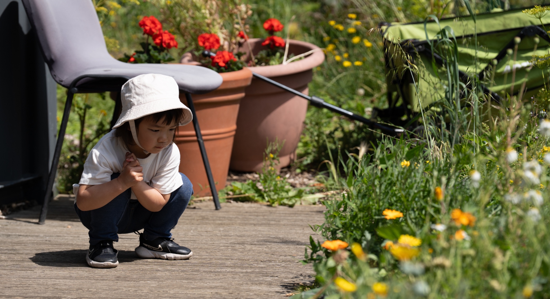 A young girl in a white hat crouches down on decking to observe the lush green and yellow plants growing in the garden surrounds.