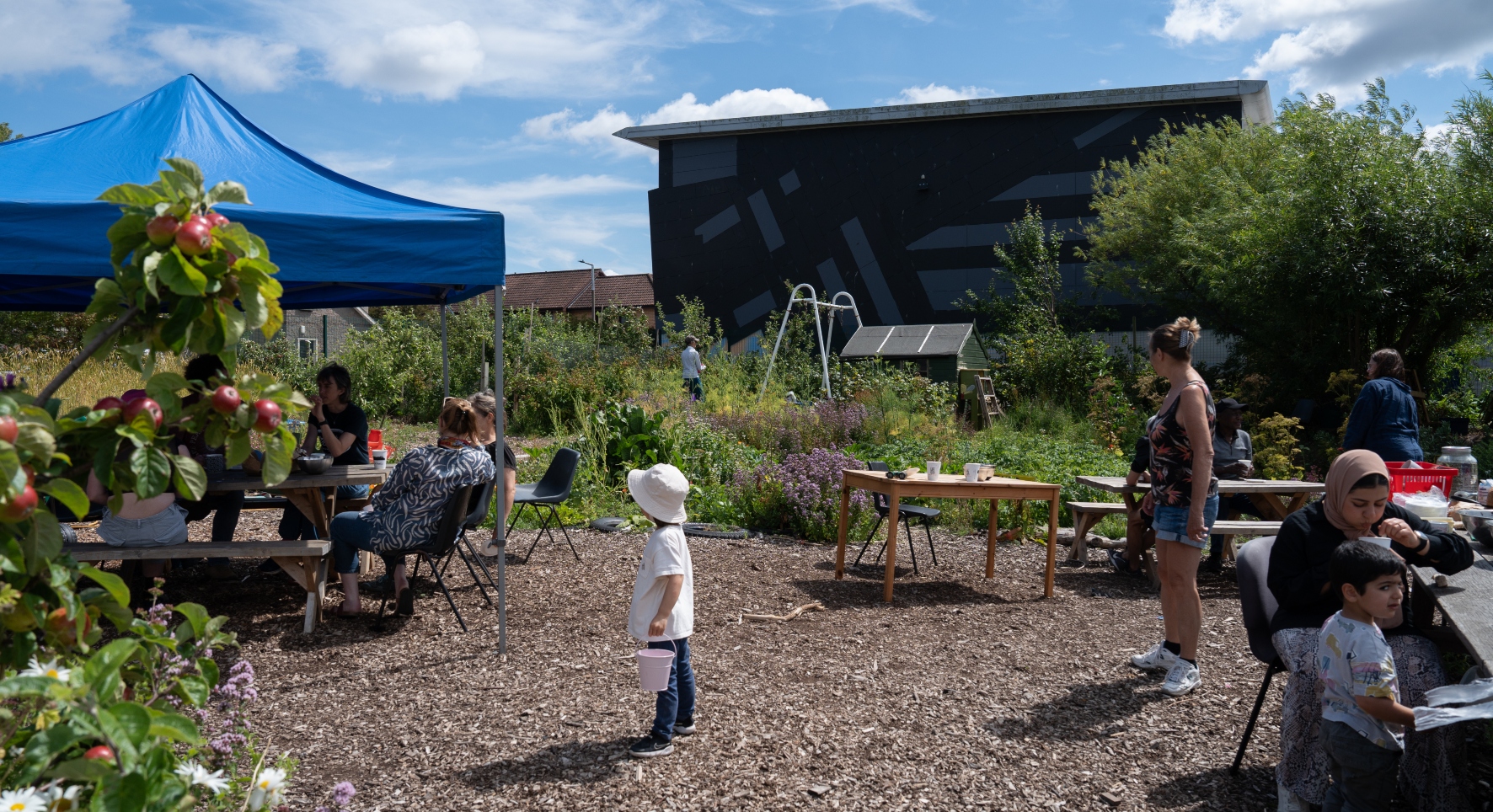 A group of people relax in an urban community garden. Some chat under the shade of a blue gazebo, others sit at sunny tables and one or two potter about doing gardening jobs.