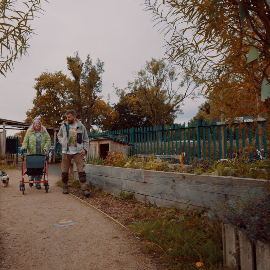 A man with a zip fleece and a lady with a green and yellow coat walk side by side along a wintry garden path. She holds a small terrier on a lead.