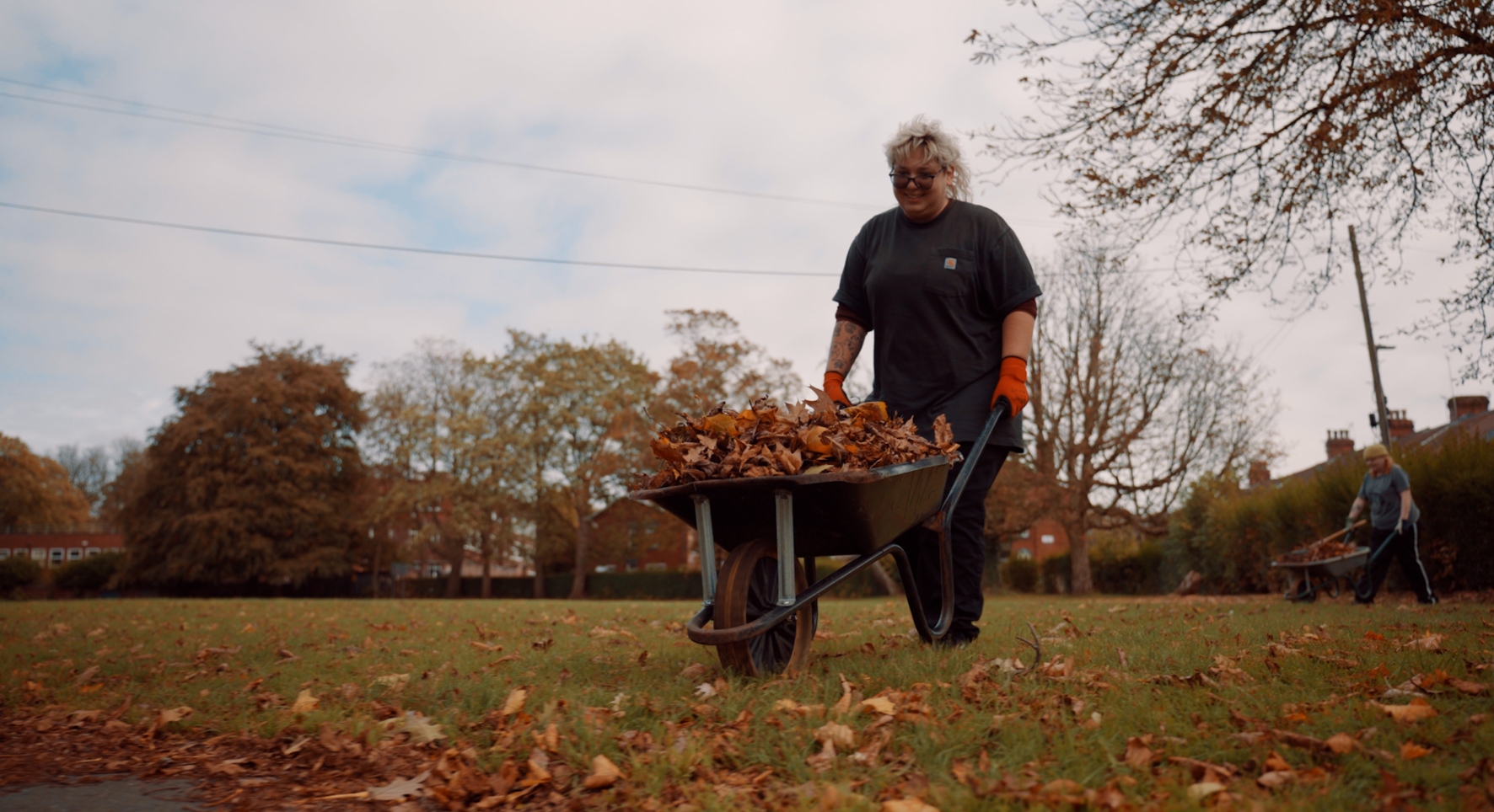 A lady in black t-shirt and trousers pushes a wheelbarrow piled high with autumn leaves over a leafy field.