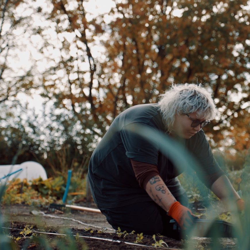 A lady kneels down at the end of a flower bed with a tray of seedlings. She wears sunglasses, bright orange gardening gloves and dark top.