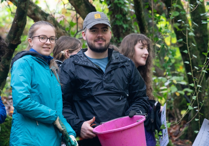 A group of teenagers stand in a dense forest setting. A girl and boy can be seen in the foreground, he carries a large pink bucket.