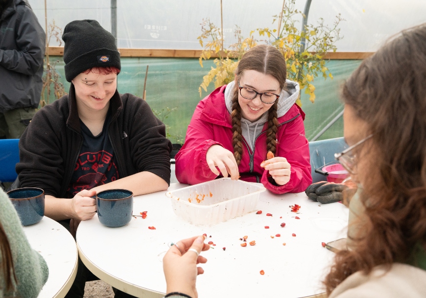 Two teenagers sit at a table. One holds a cup of tea or coffee and the other is sorting some acorns into a plastic tray. A tree sapling grows behind them.