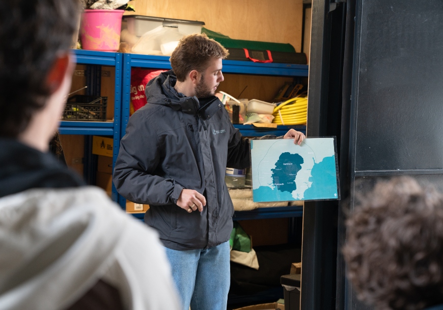 A young man holds up a map, standing in front of a series of shelves with outdoors equipment.