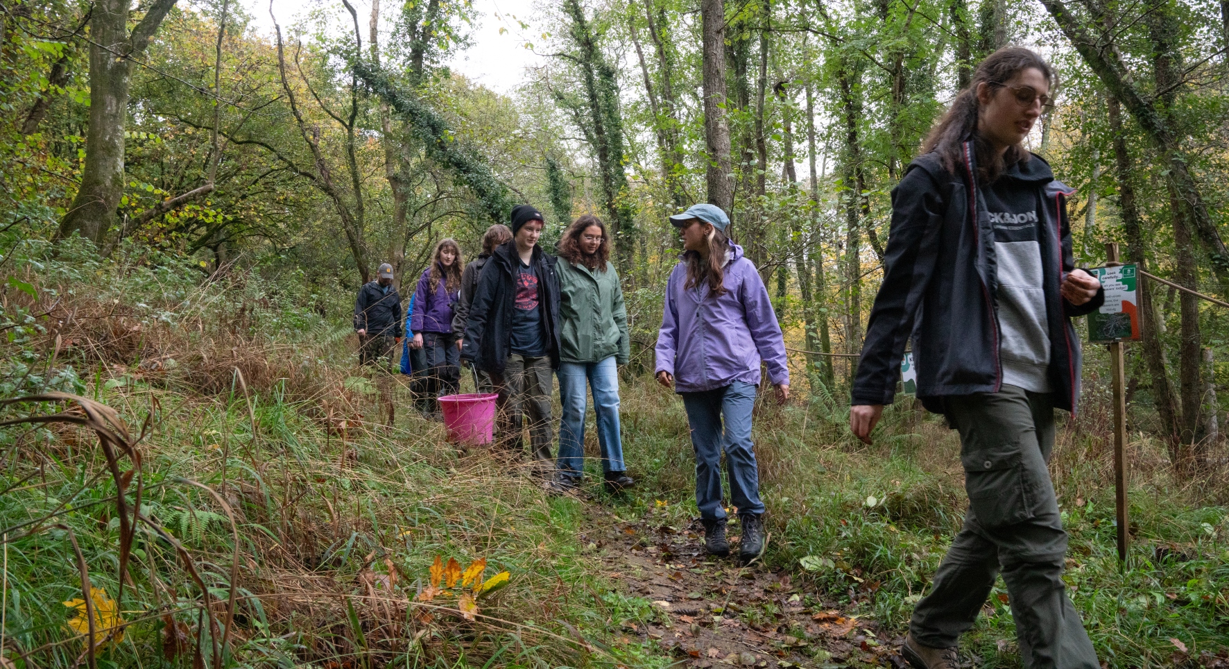 A group of young people walks in a loose line along a forest track. They're in wintry clothing and one carries a large pink bucket.