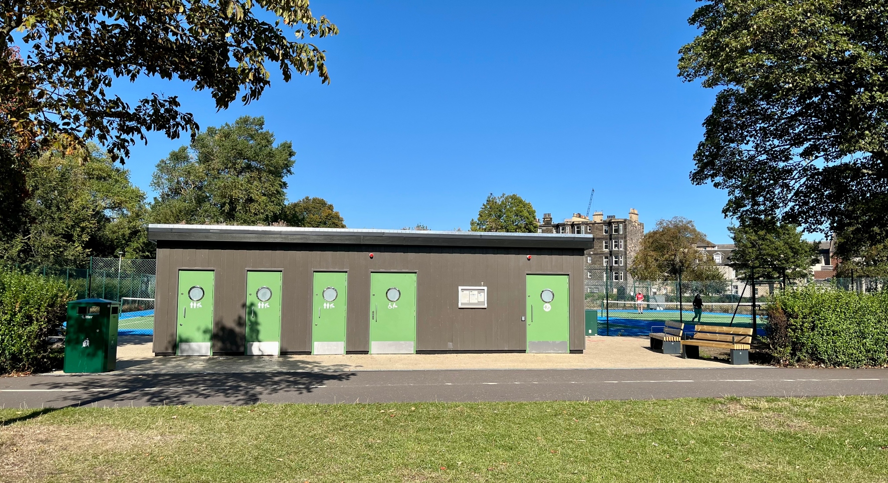 A block of toilets in a public park. They are brown-cladded with green doors. The park has an autumnal feel with fallen leaves.