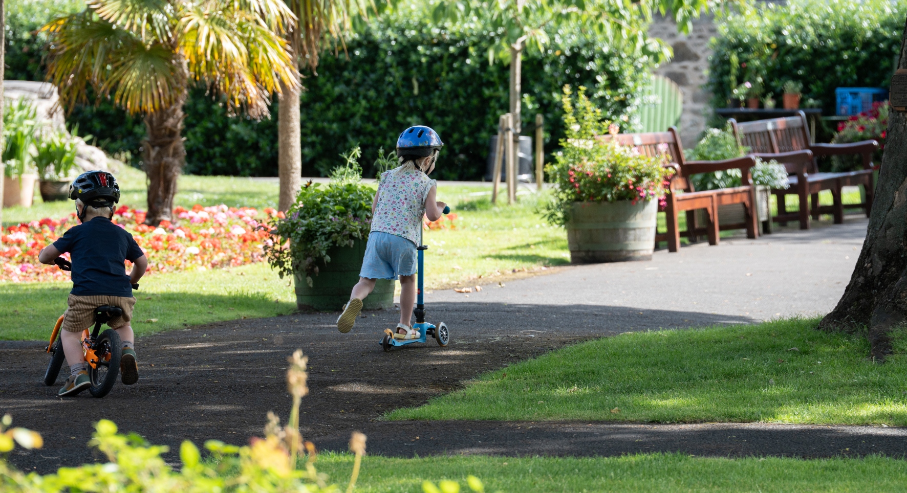 Two young children play in a sunny park on a summer's day. One rides a scooter whilst the other wheels on his balance bike. 