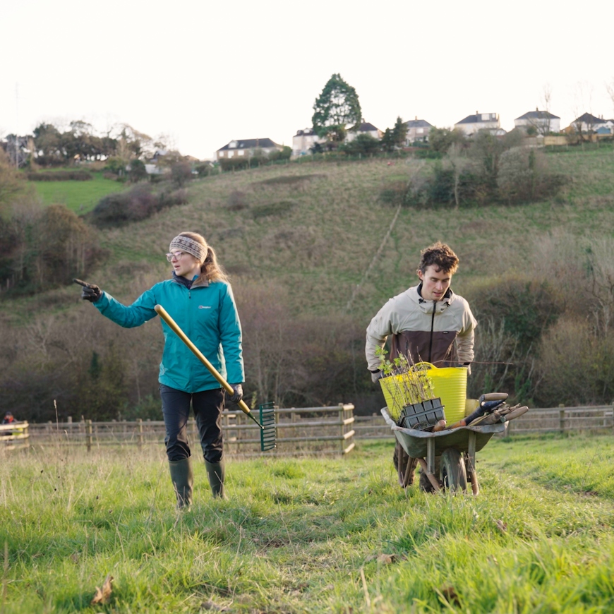 A young woman and young man walk uphill through a wintry field. She carries a rake and he's pushing a wheelbarrow full of gardening tools. Trees and the outskirts of an urban housing estate rise behind them.