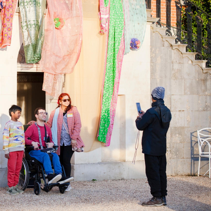 A group of three pose for a photo against a wall decorated with beautiful fabric hangings. They're a mixed group including a young boy and a man in a wheelchair.