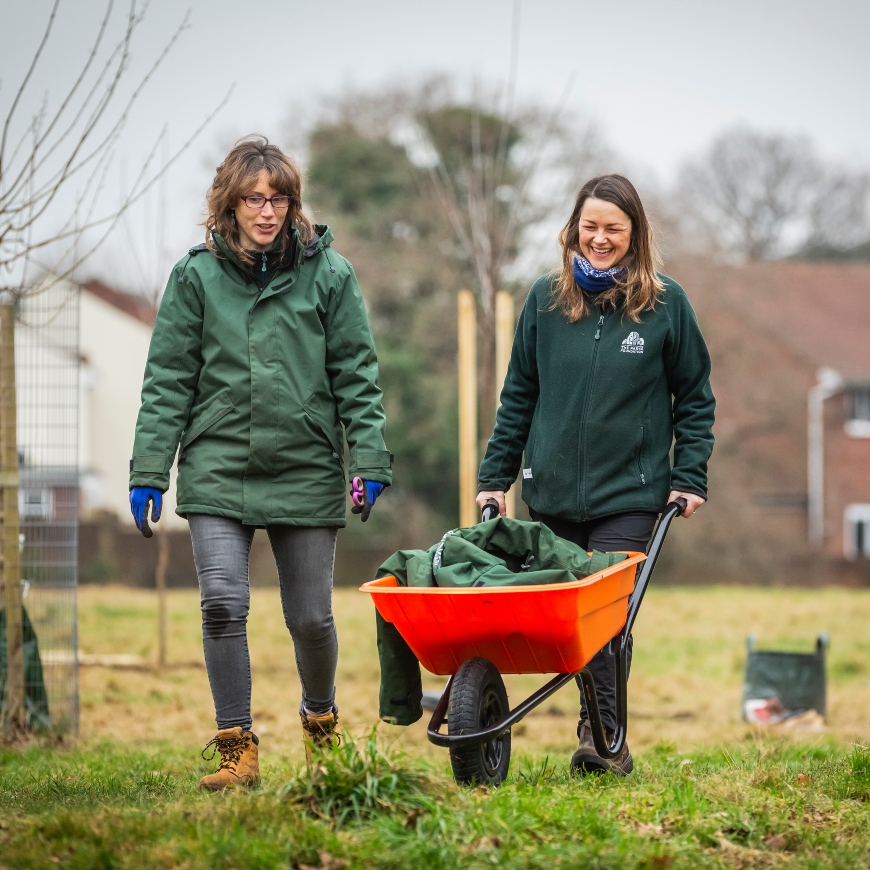 Two women, both in green, smile as they walk through a park field on a wintry day. They push a bright orange wheelbarrow, in stark contrast to the grey sky and spindly trees behind.