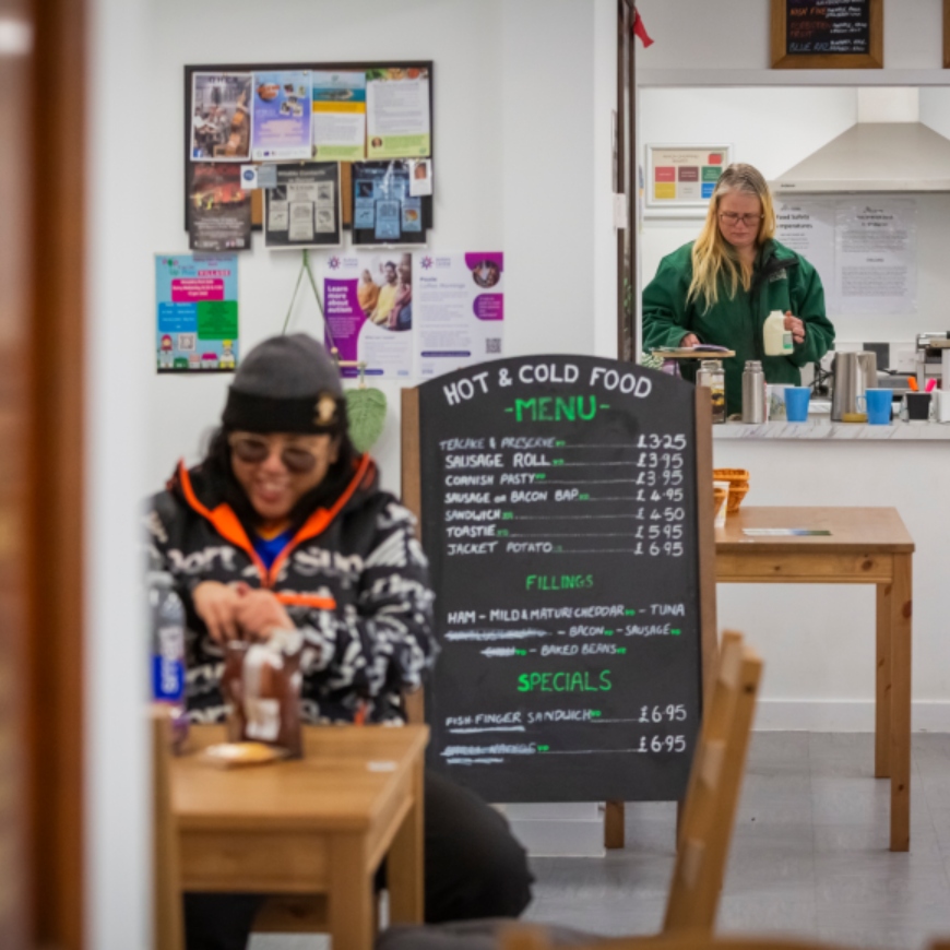 A lady in a black hat and raincoat tucks into a drink and snack at a cafe table. Behind her is a lady in a green fleece, preparing tea at a serving hatch.