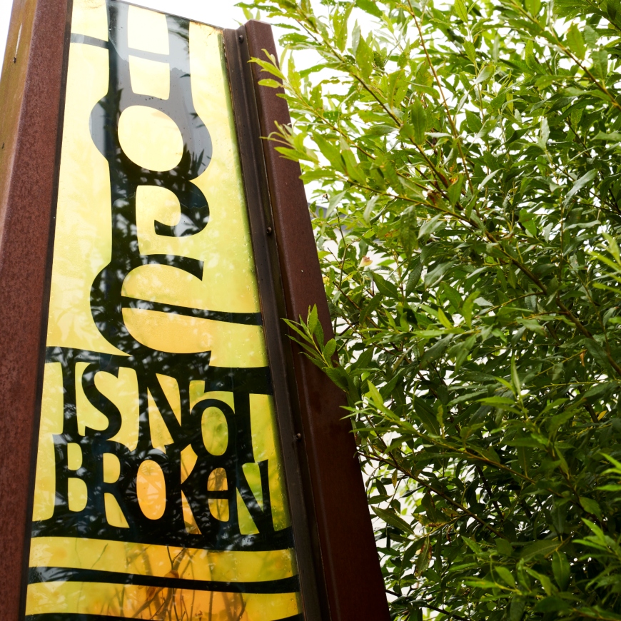 A large, golden sign looms along the left hand side of the image, with wooden frame and black lettering that says 'Hope is not broken'. Trees filter over the sign on the right.