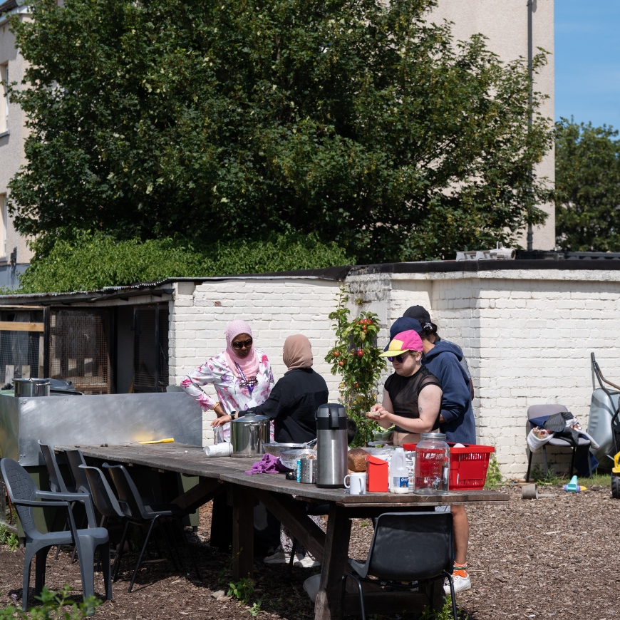 A group of people from diverse cultures share a meal together in a community garden. It looks like a relaxed, chatty event with everyone having a role to play.