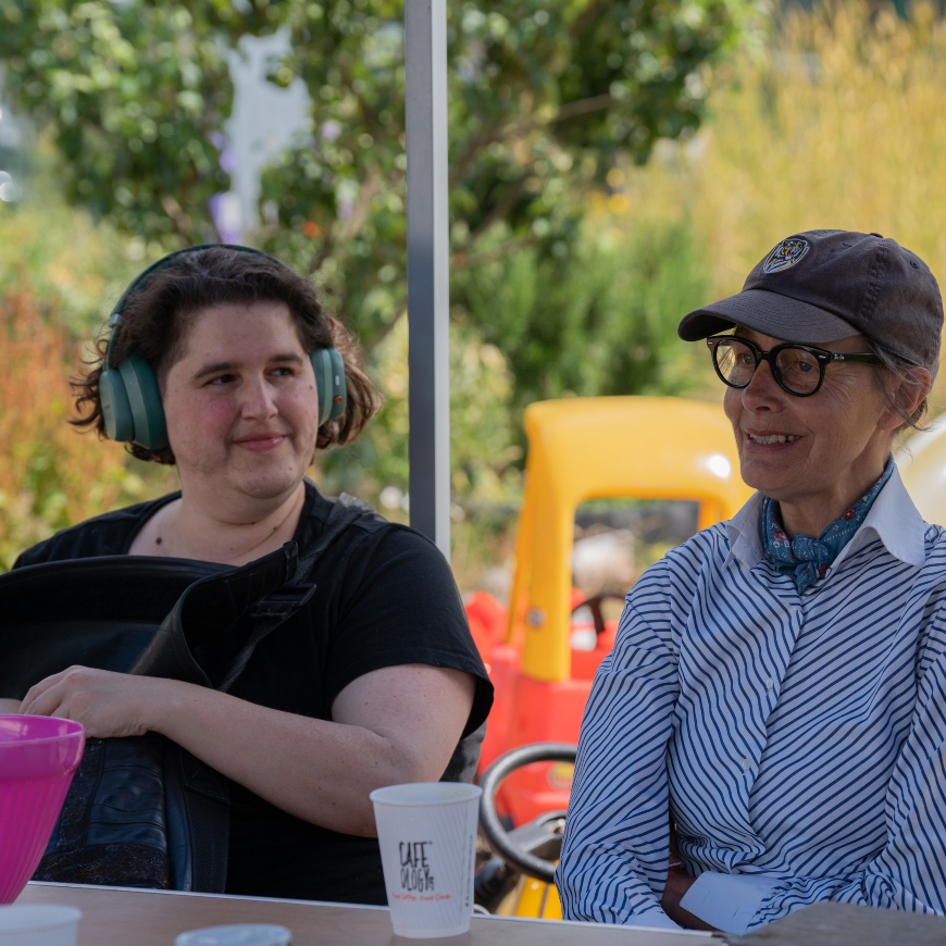 Two women sit at a bench, relaxing over a cup of tea. One wears a sunhat and the other has green headphones. It's a lovely summers day with lush greenery behind them.