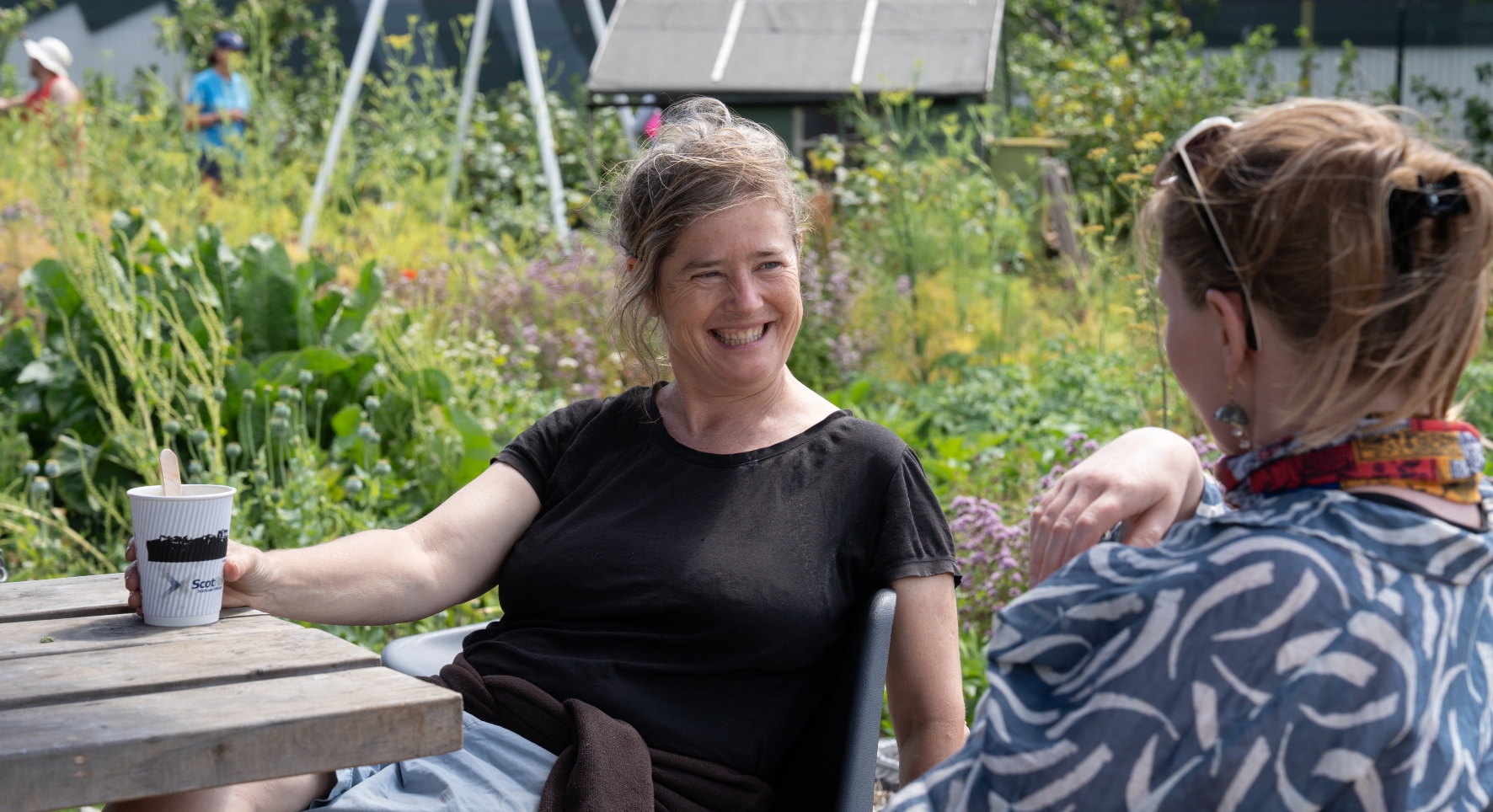Two women laugh together over a cup of tea. They're sitting at a wooden bench, with a lush community garden behind them.