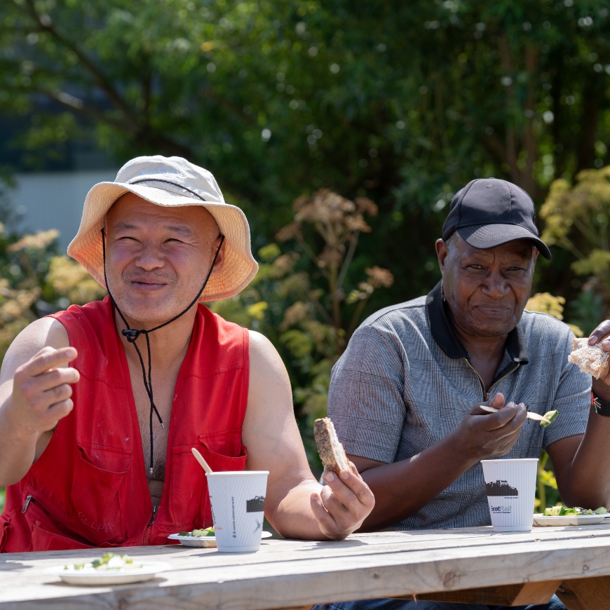A couple of men sit at a wooden bench on a summers day. They're tucking into tea and biscuits and look to be sharing a joke.