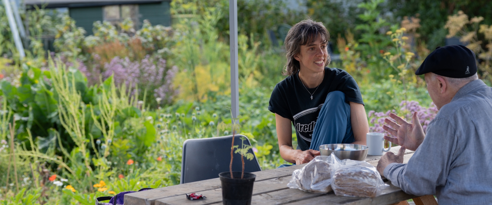 Two people sit and chat on a summers day. They're at a bench with tea and what looks like locally-grown produce, most likely from the lush garden behind them.