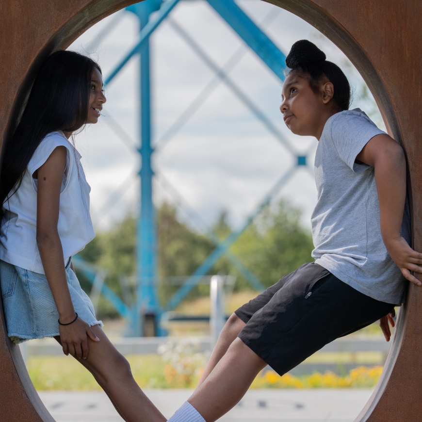 Two children stand in a large, metal letter 'O' in the middle of an urban park.