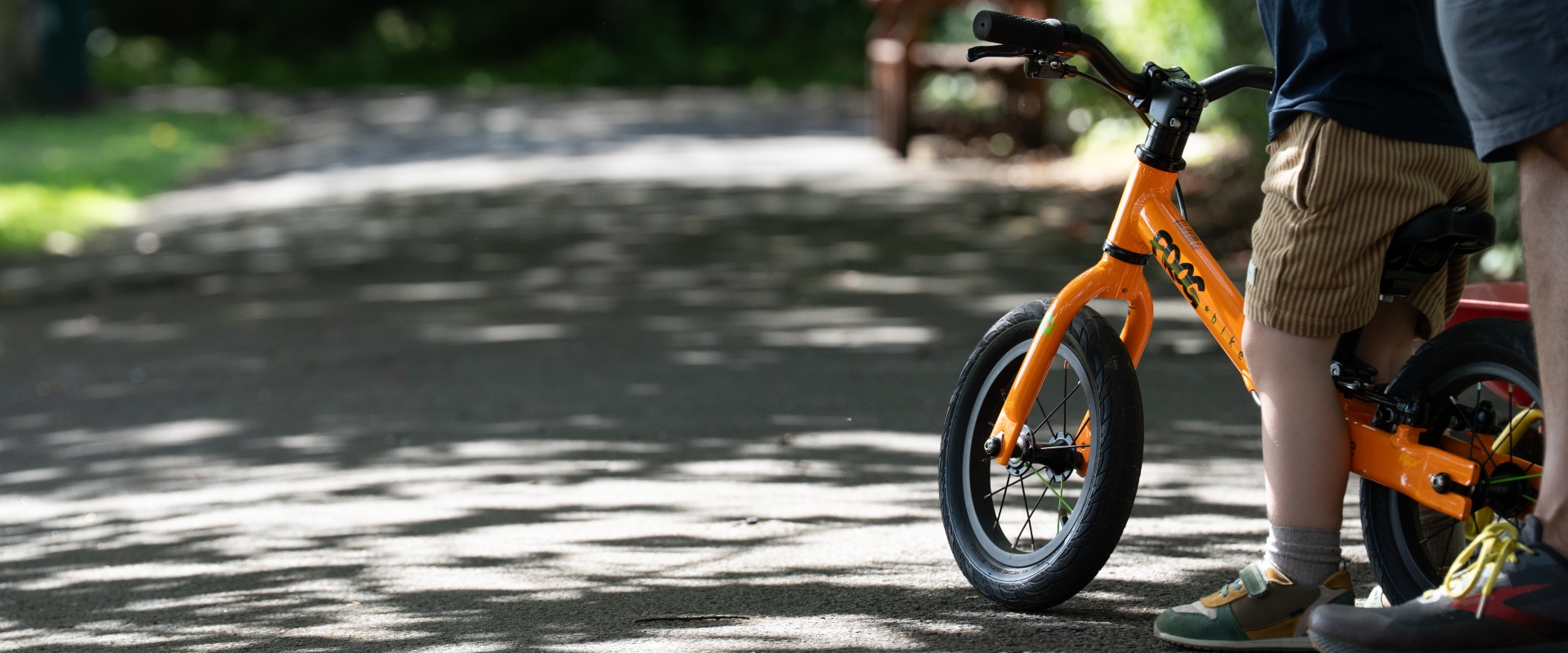 A close-up of a bright orange balance bike, complete with the legs and feet of its rider, on a sunlit path