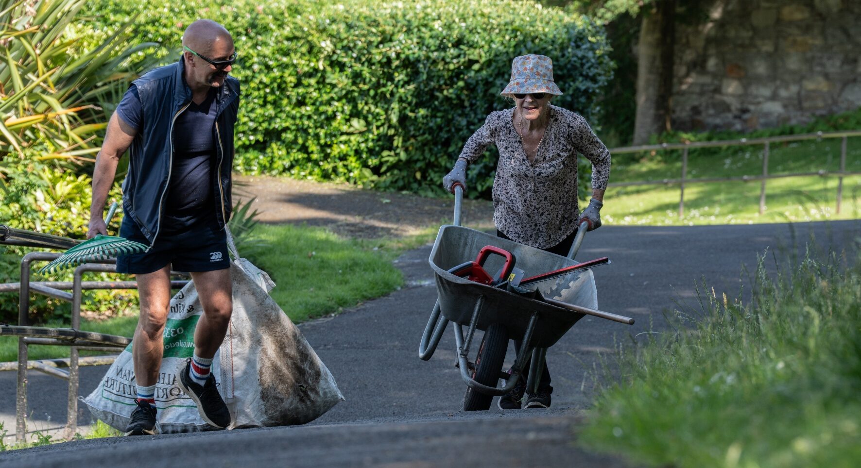 Two people hard at work in a large garden. A lady pushes a wheelbarrow, whilst the man drags a large sack in one hand and carries a rake in the other. Despite the hard graft, they look cheery.