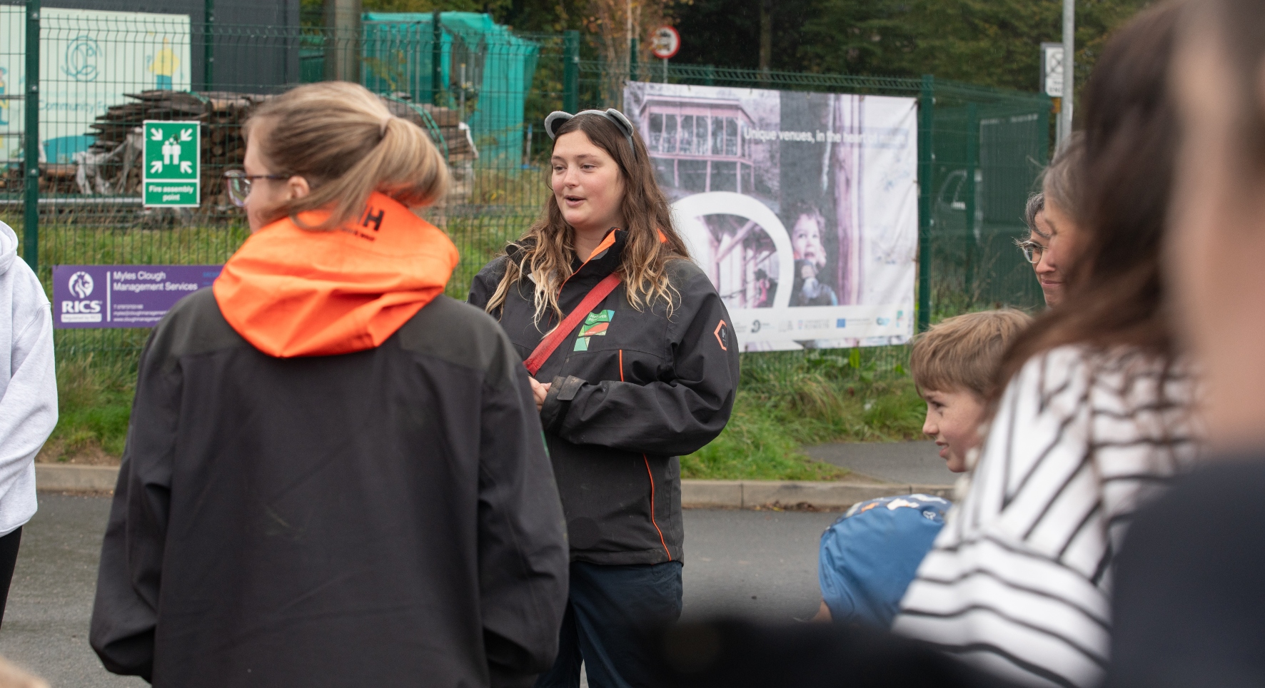 A young women wearing bear ears chats to a group of mixed-age people. They're in an urban setting and look like they're about to embark on a walk.
