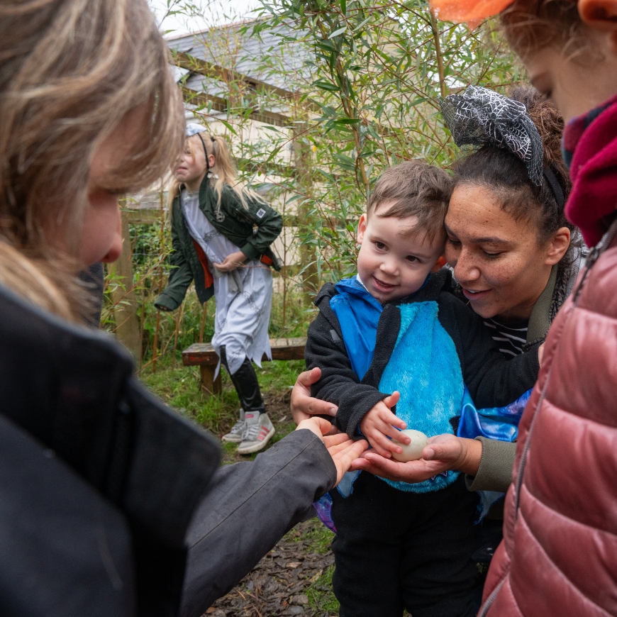 A little boy in a blue top smiles as he reaches out to touch a white chicken egg. He's being helped out by lady with a halloween-themed headpiece
