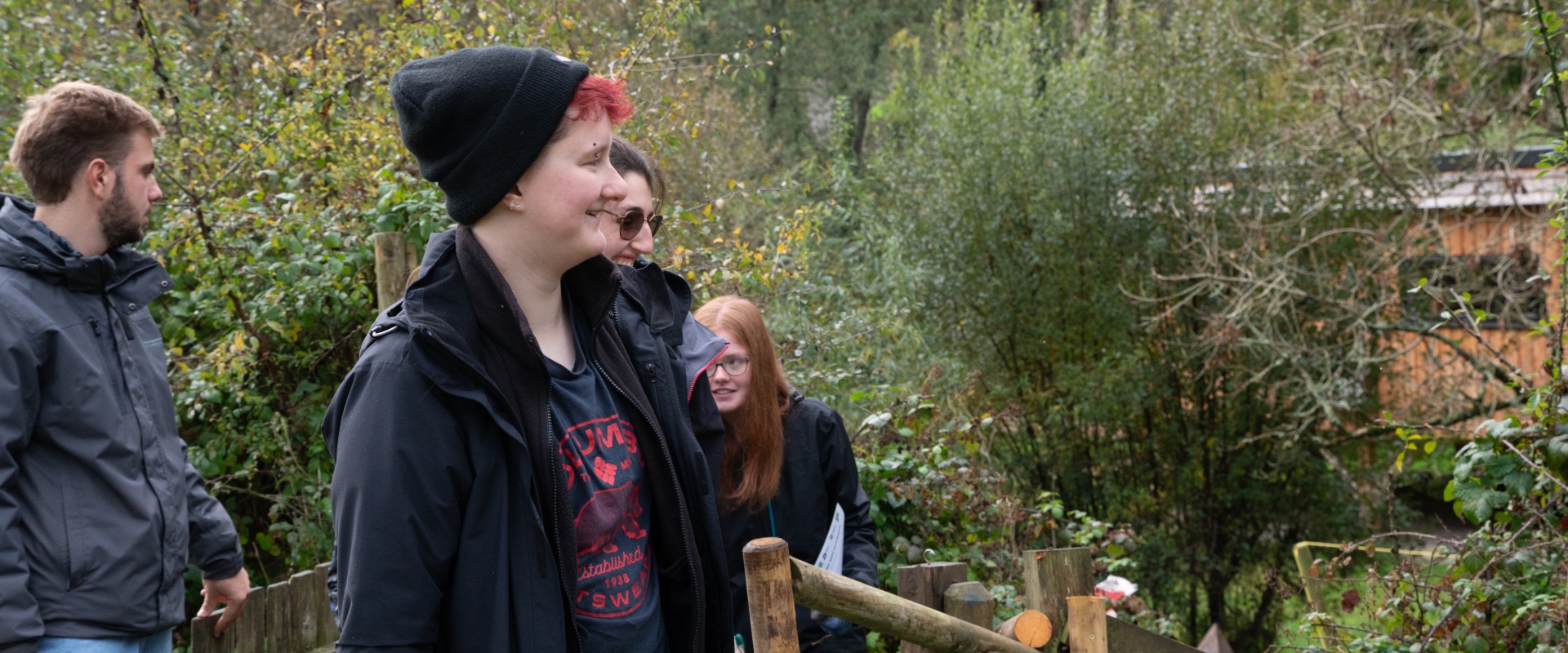 A young person with black beanie smiles as she looks over a forest area. There's a group of people walking up behind her and lots of green foliage and trees.