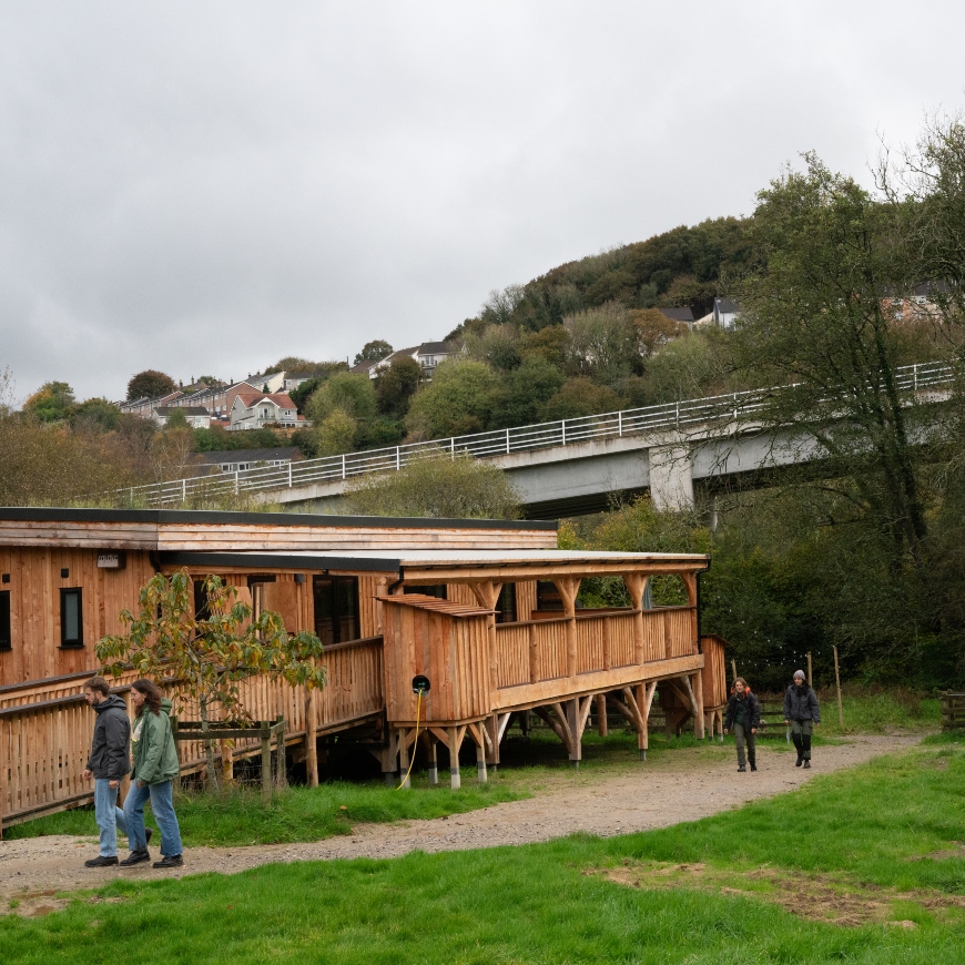Young people walk in front of a large wooden building on stilts. It's situated in a city woodland, with trees, housing and a grey flyover cutting across the top of the image.