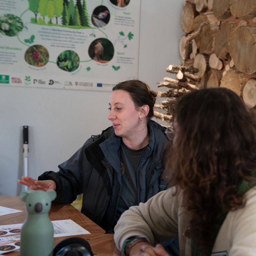 A lady sits at a desk, chatting to a person sat beside her. A stack of logs and poster with trees on it sit behind them. They look to be having a purposeful but positive chat.