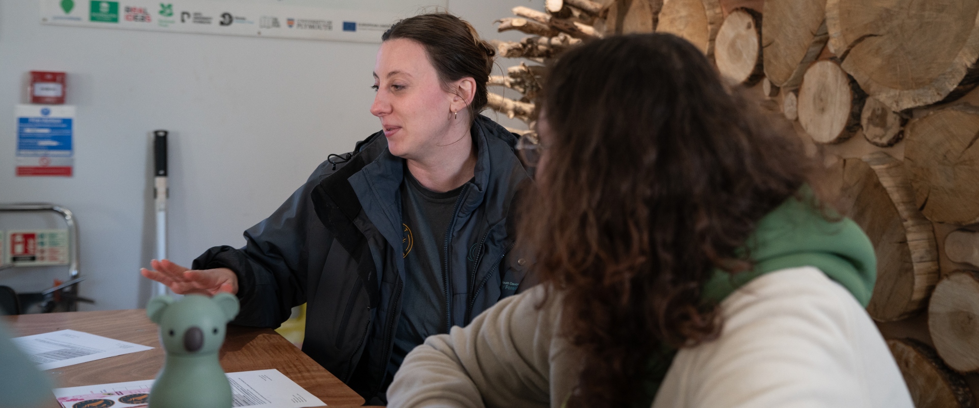A lady sits at a desk, chatting to a person sat beside her. A stack of logs and poster with trees on it sit behind them. They look to be having a purposeful but positive chat.