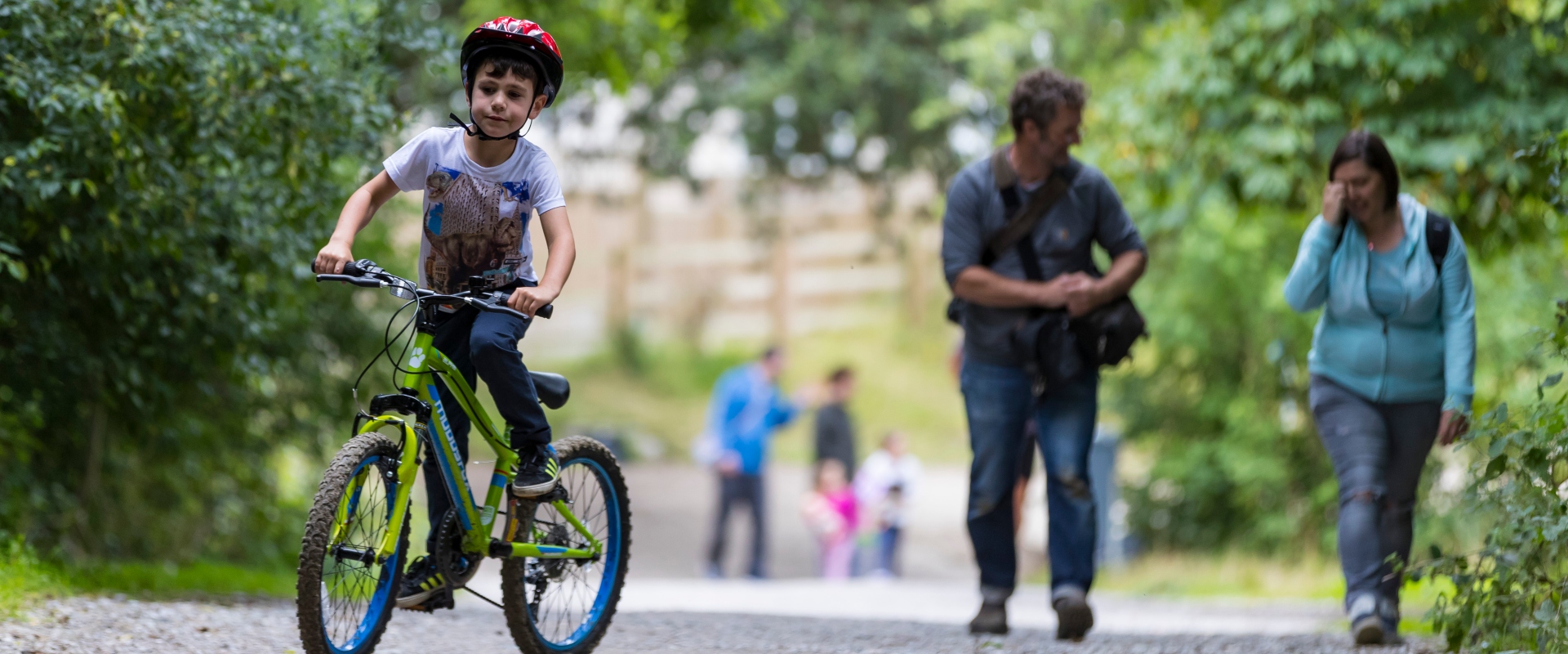 A young boy rides his bike along a path on a summers day. Two grown-ups walk just behind him. They're surrounded by lush hedges and trees, but look to be in a city with large buildings in the background.