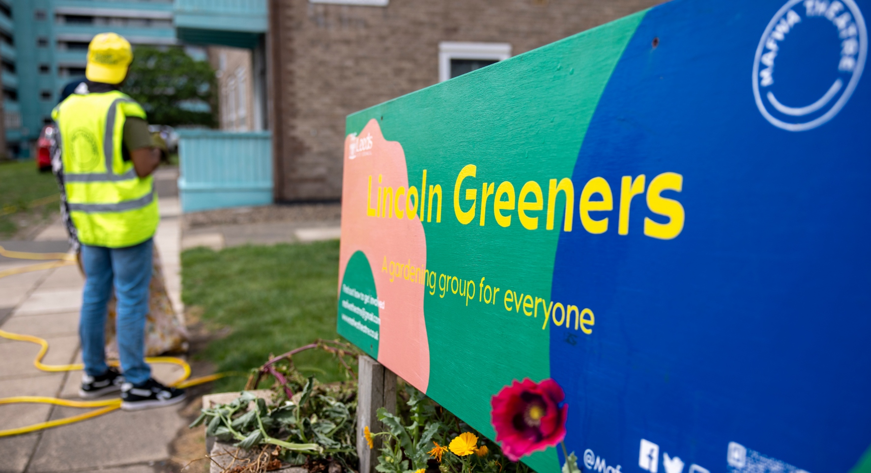 A large, bright sign says 'Lincoln Greeners' along the right hand side of the image. To the left, a man stands with his back to the camera wearing high-vis yellow. They're in a city garden area, with tall flats and lawns.
