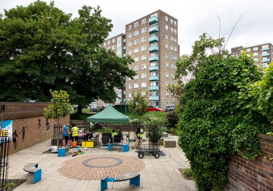 Roxby Community Garden in the Lincoln Green area of Leeds. There's green gazebo shading people busily tending to plants, whilst others pause for a break over coffee. Tall residential buildings surround the garden.