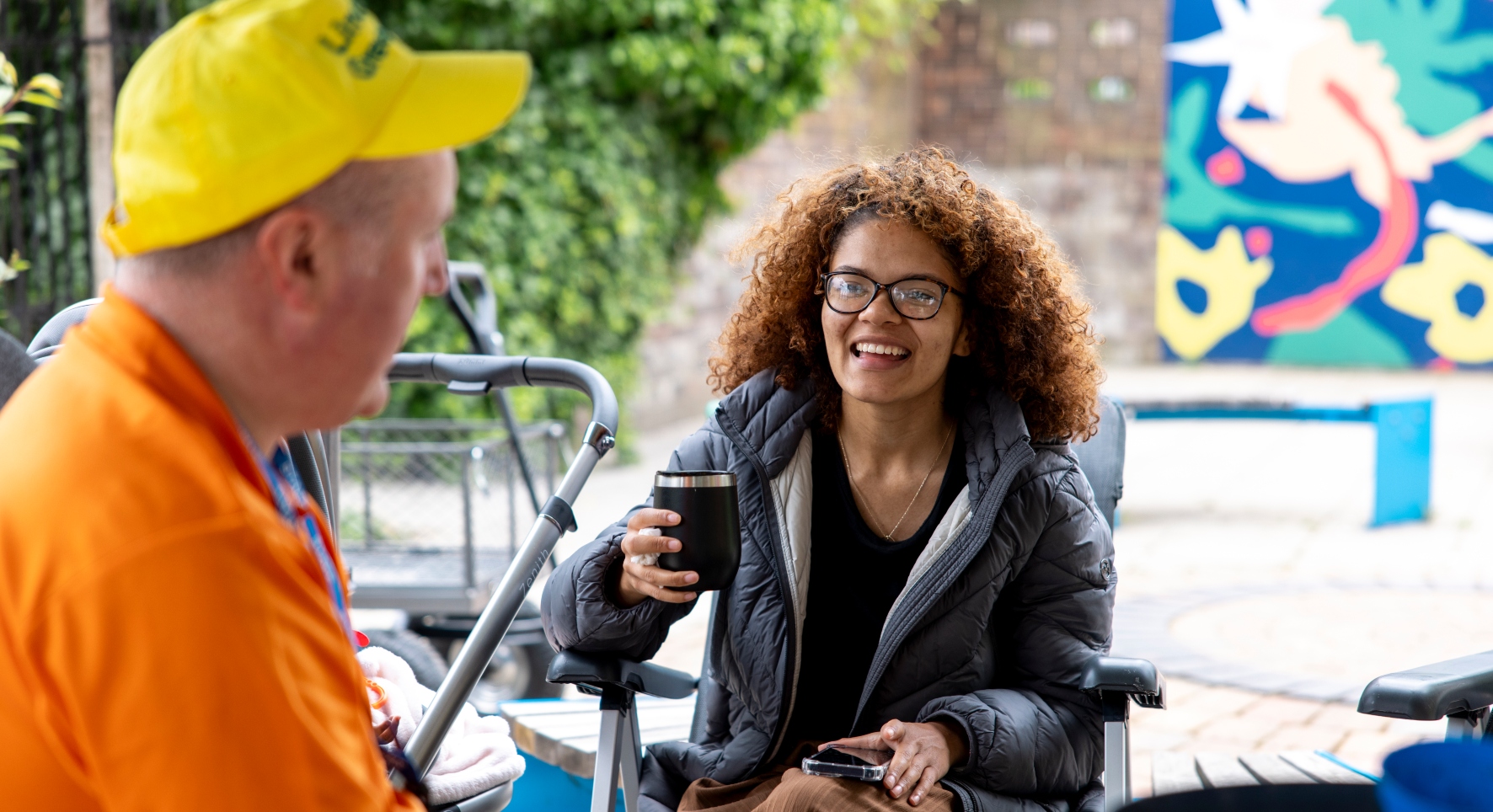 Two people sit and chat over hot drink. They look to be in an urban garden area, with paving and bright artwork along with trees and shrubs behind them.