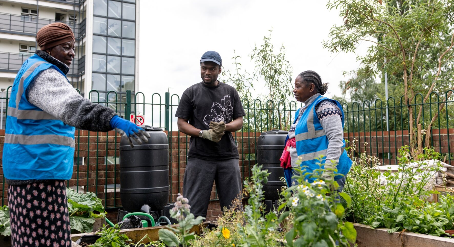 Three people stand in the corner of a fenced area. There are raised flower beds in the foreground and tall buildings rise behind. Two of the people wear blue high-vis jackets and the third is ready for action in gardening gloves.