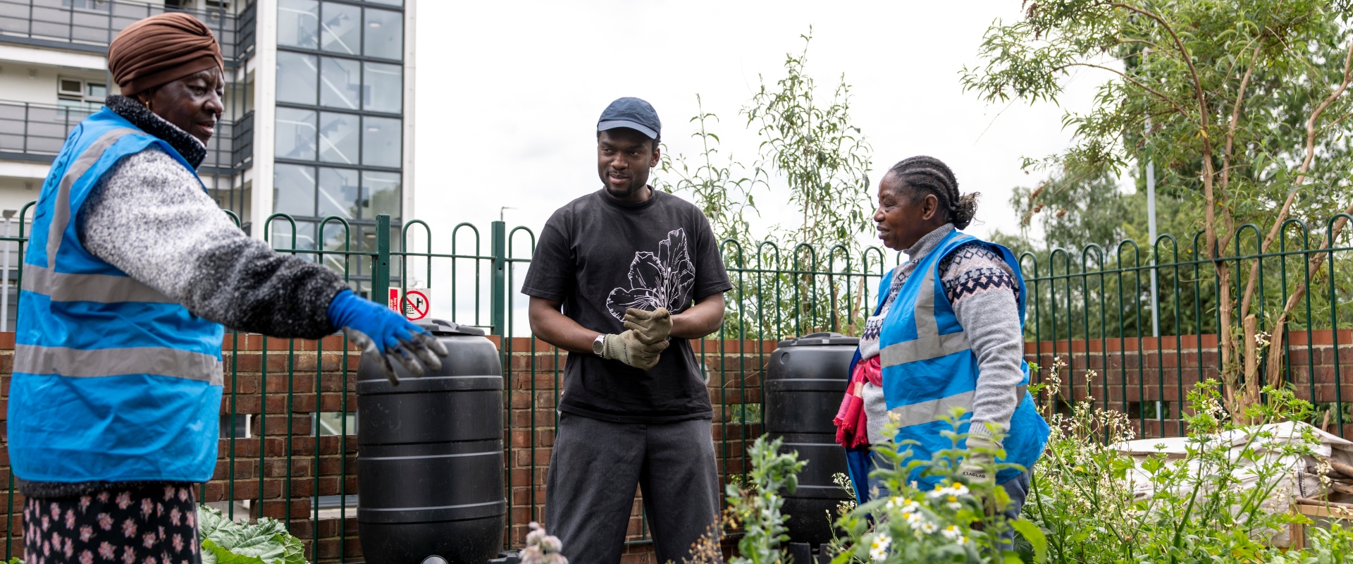 Three people stand in an area of greenery, engaged in conversation. The two women are wearing blue high-vis jackets, whilst the man stands in the centre with a black cap and t-shirt. They look to be in an urban garden, with a low wall, railings and building behind them.