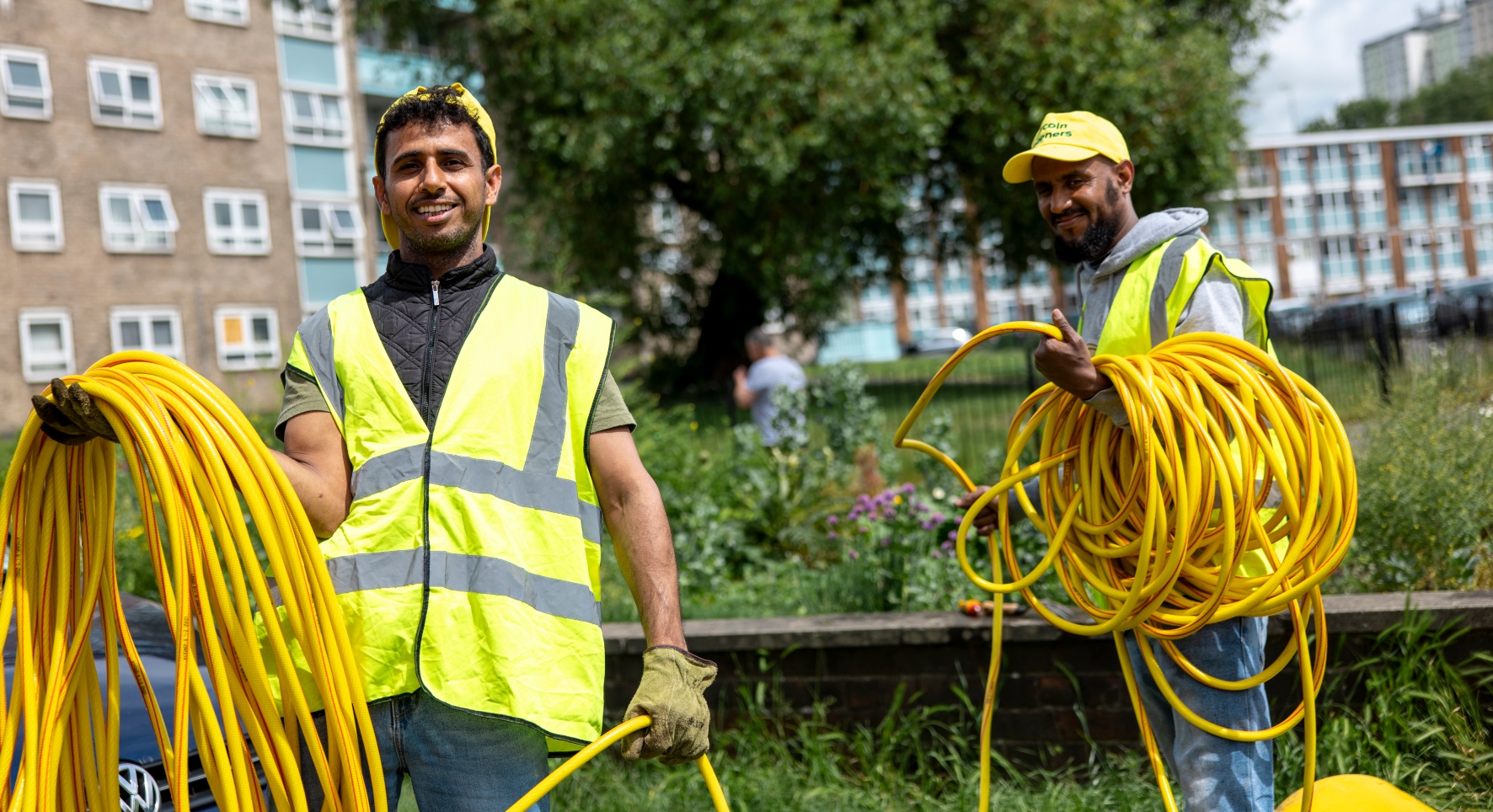Two men stand in an area of greenery and flowers. They wear yellow high-vis jackets, yellow sunhats and each carry a yellow hose wound around their arms. The lush scene is juxtaposed by big buildings and blocks of flats, suggesting they're in an urban area.