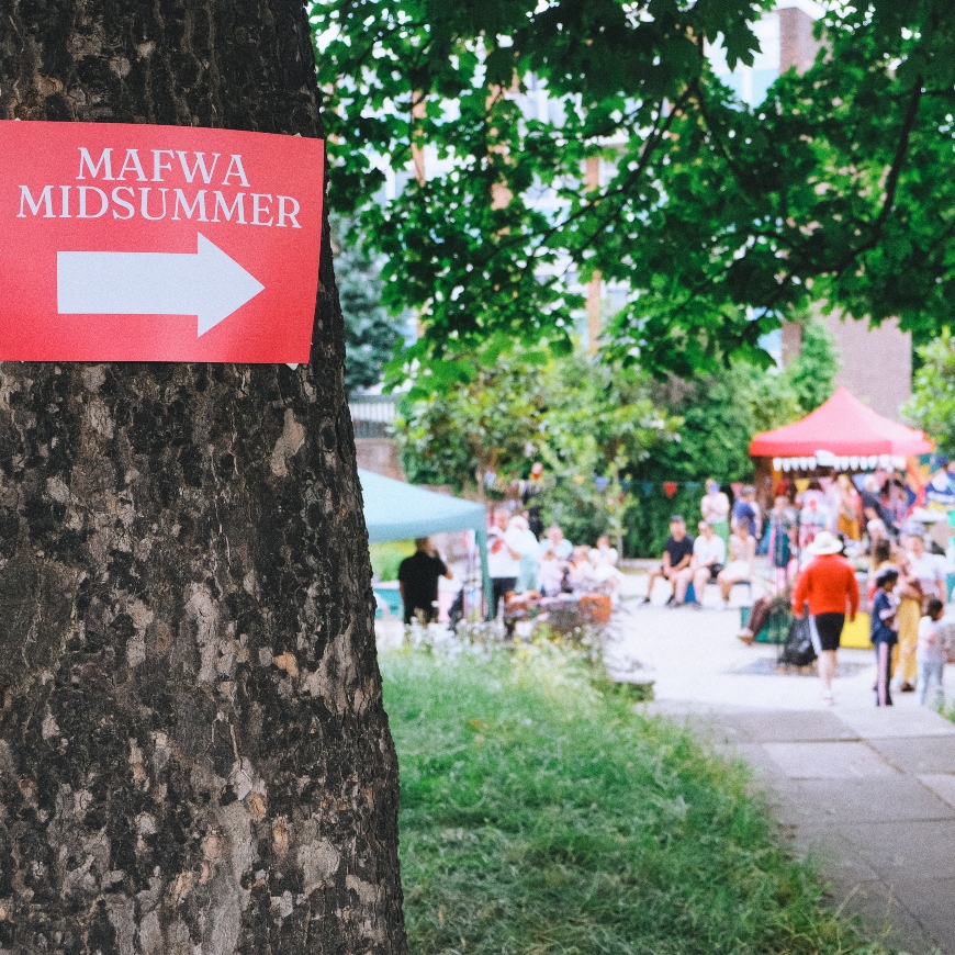 A tree trunk takes up the left hand side of the image, with a red 'Mafwa Midsummer sign'. A white arrow draws the eye to the right of the image, where a crowd in an urban garden looks to be celebrating in the sunshine with flags and gazebos.