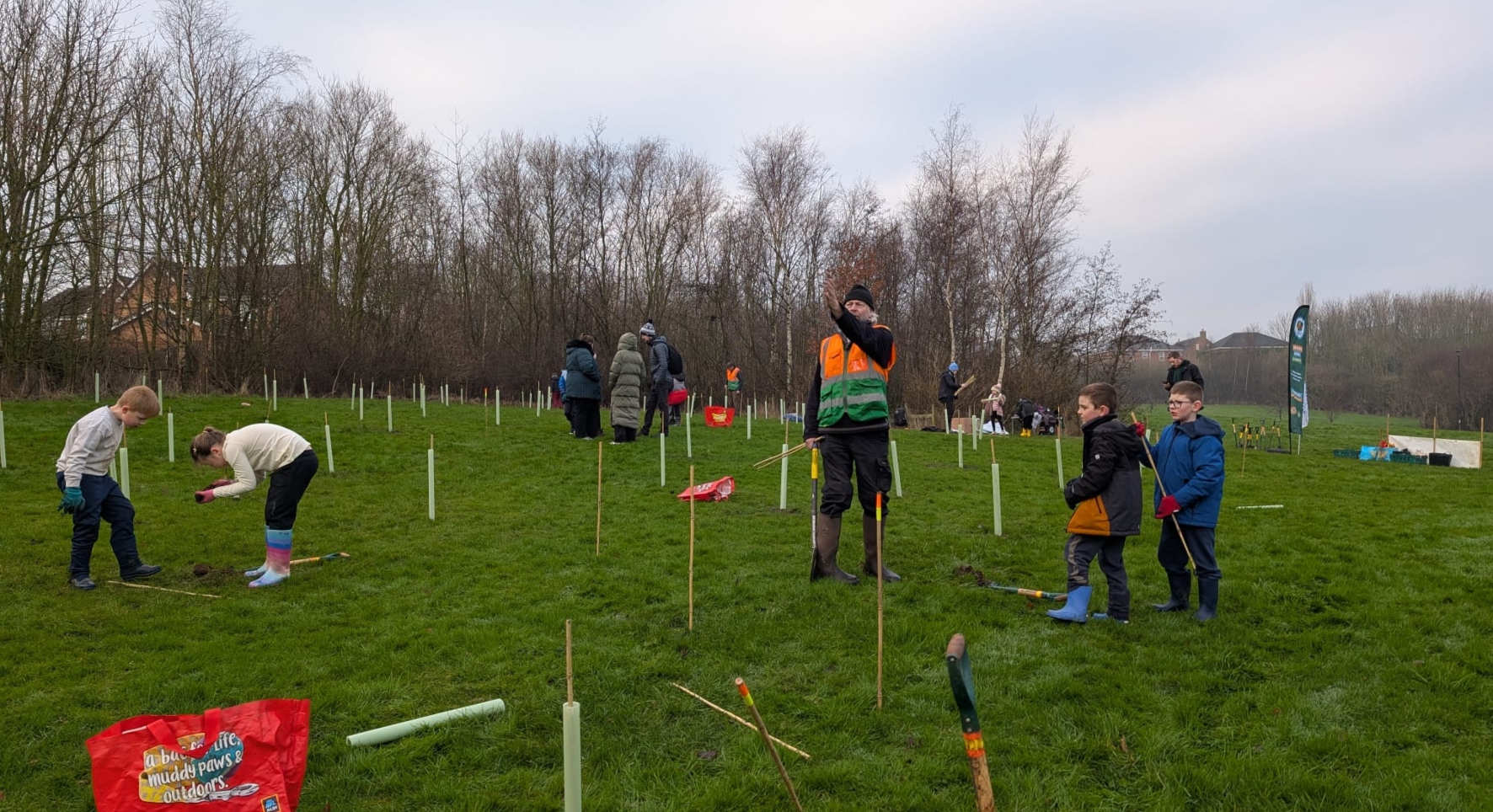 People plant young trees together in a field. There's a mix of ages, from kids to grown-ups. A man in high-vis stands in the middle, pointing off to the distance.